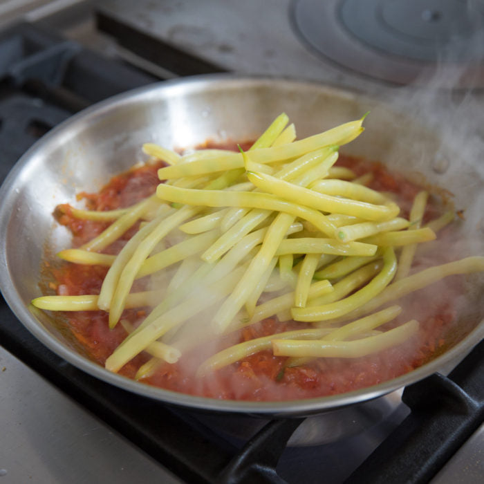 Yellow wax beans being braised in tomato sauce in a stainless-steel skillet on a gas stove