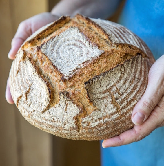 Hands holding a round whole-grain einkorn boule with scored crust and a flour-dusted spiral pattern