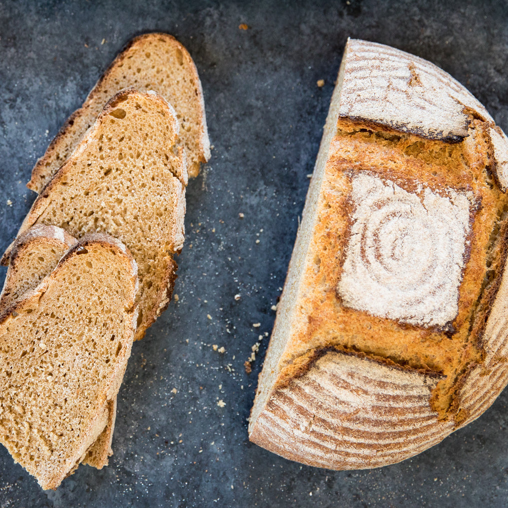 Sliced organic einkorn bread loaf on a dark stone surface showing airy crumb and flour-dusted crust