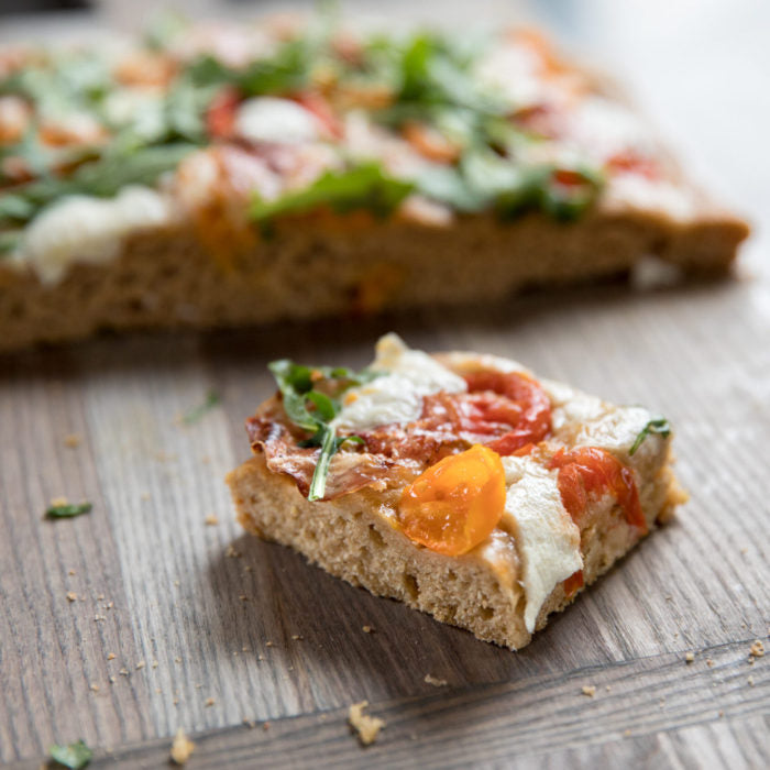 Close-up of an Einkorn whole wheat focaccia slice topped with cherry tomatoes, melted mozzarella, and arugula
