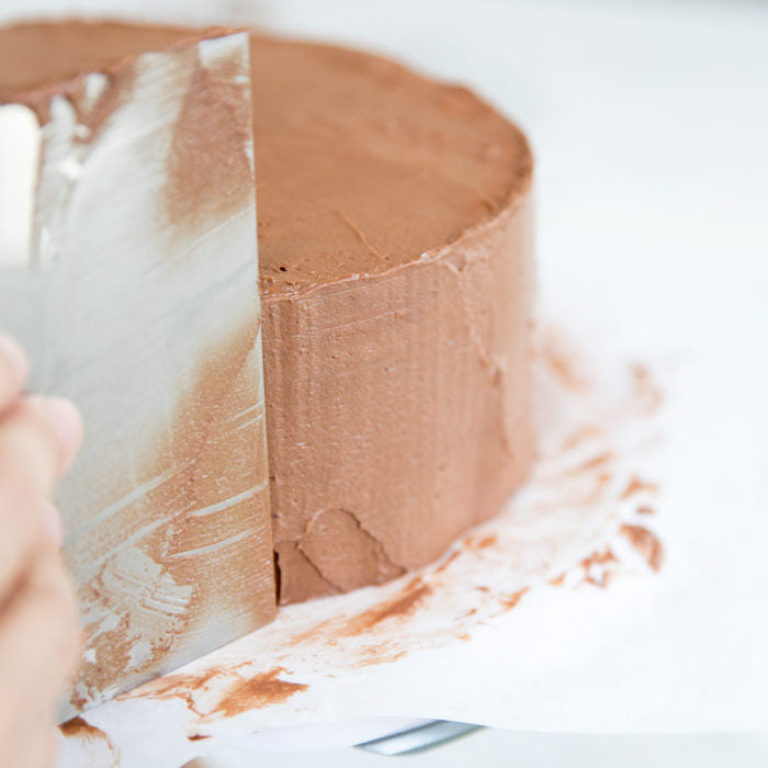 Three-quarter view of an Einkorn yellow cake being frosted with chocolate buttercream, spatula smoothing frosting on white parchment