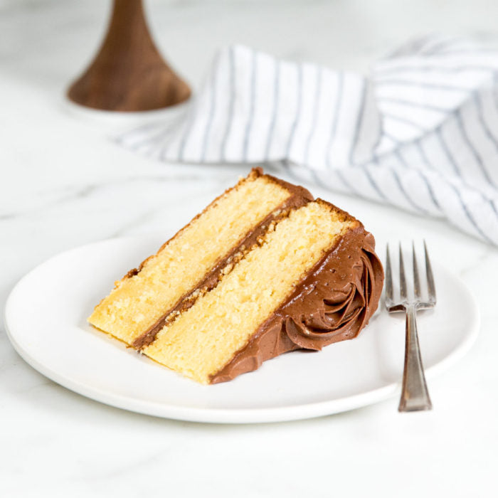 Three-quarter view of a slice of yellow Einkorn cake with chocolate buttercream showcasing two layers on a white plate with a fork and striped napkin