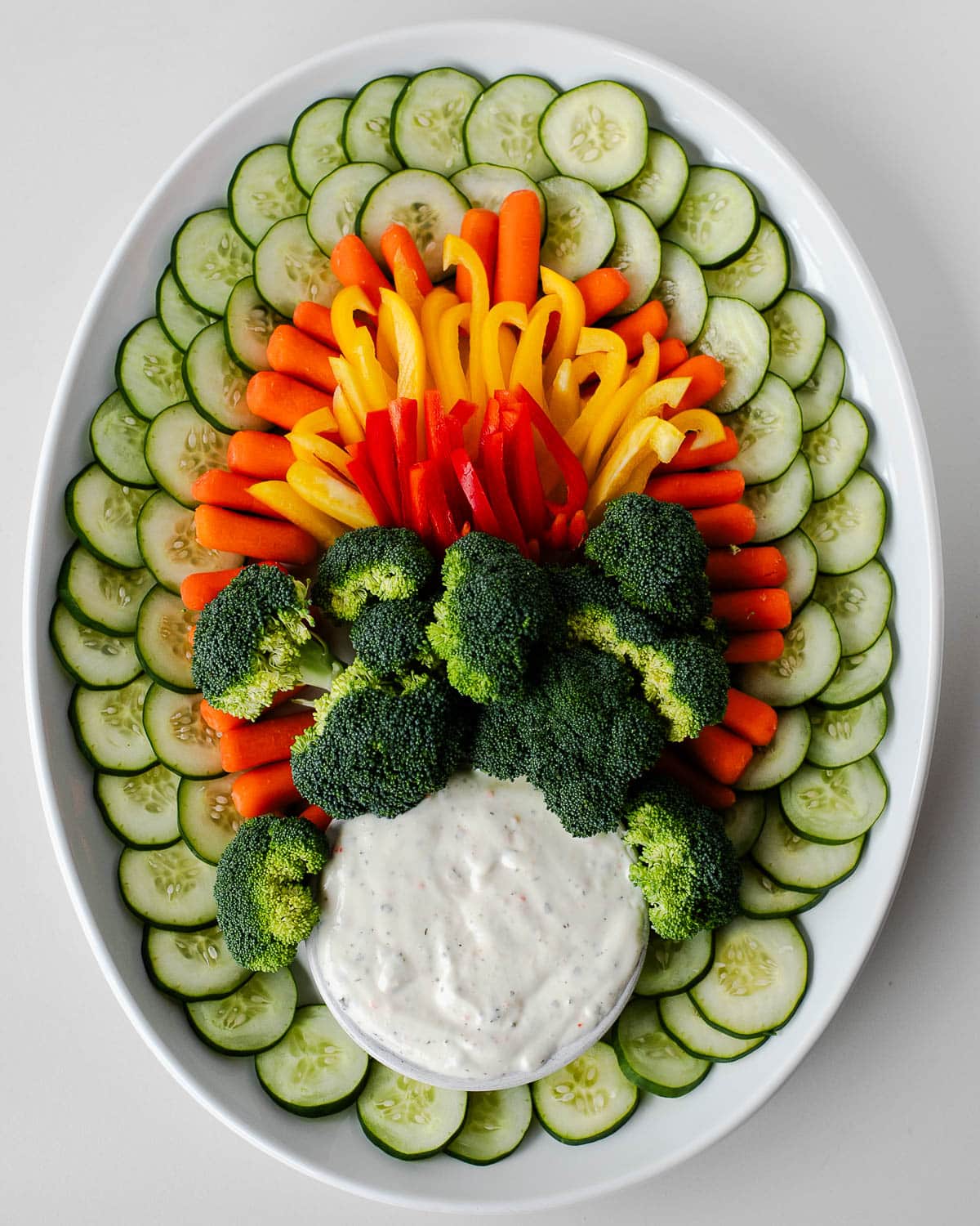 olorful layers of cucumbers, baby carrots, yellow peppers, red peppers, and broccoli arranged in a fan shape behind a bowl of ranch dip to create turkey feathers on a veggie platter.