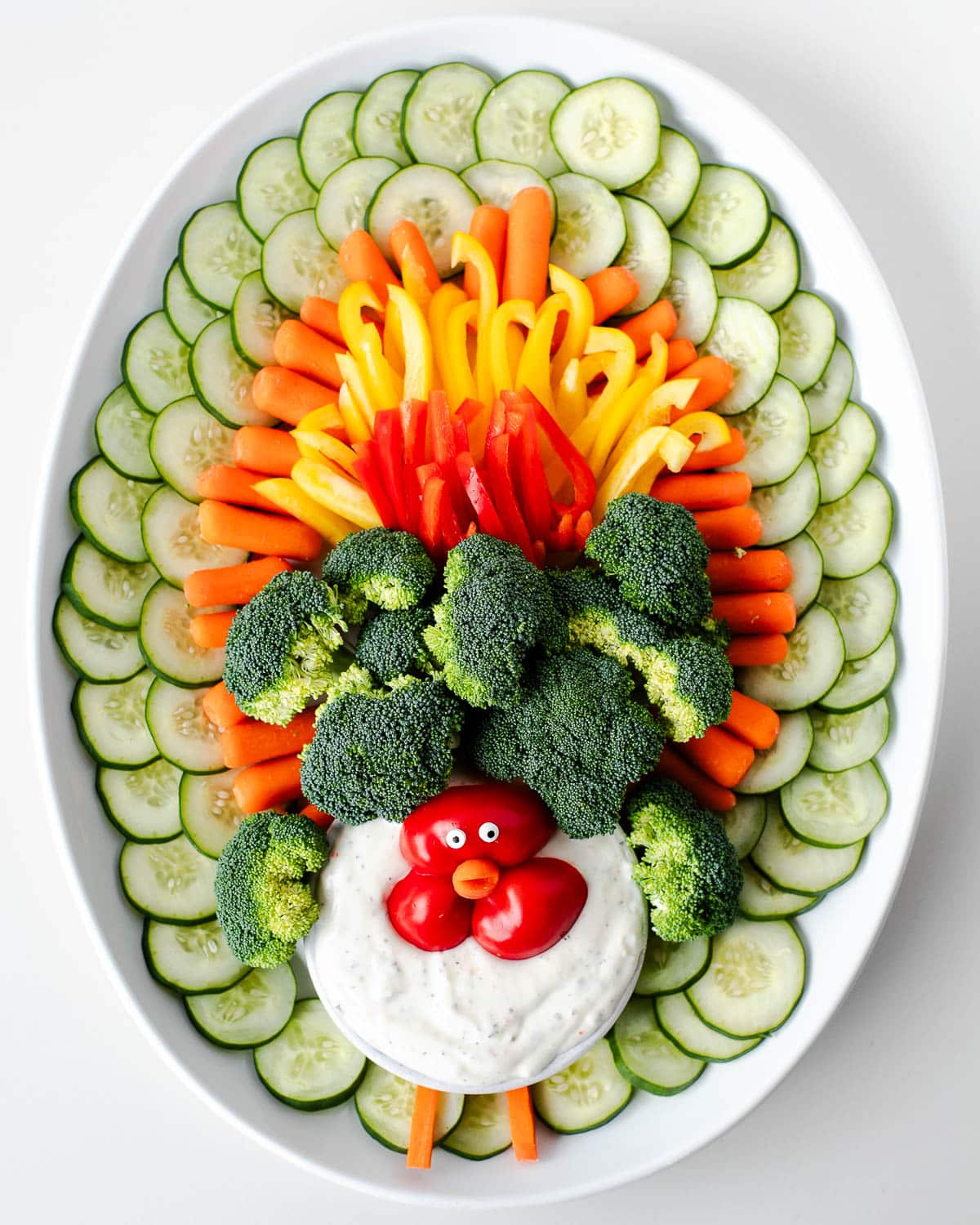 Completed turkey veggie tray with cucumber slices, carrots, bell peppers, broccoli, and a red bell pepper turkey face with candy eyes arranged around a bowl of dip on a white platter.