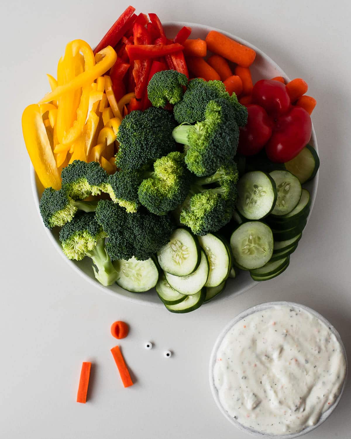 Bowl of sliced cucumbers, bell peppers, broccoli, carrots, and small decorations next to a bowl of creamy dip, showing the ingredients needed to assemble a turkey veggie tray.