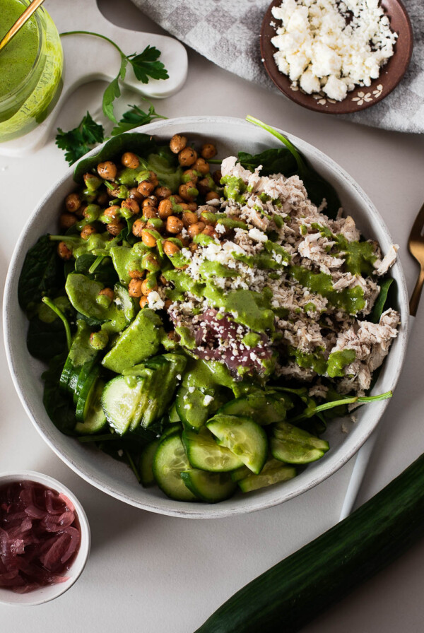 Overhead green goddess chicken bowl with spinach, cucumbers, shredded chicken, and roasted chickpeas, drizzled with creamy herb dressing; feta and pickled onions on the side.