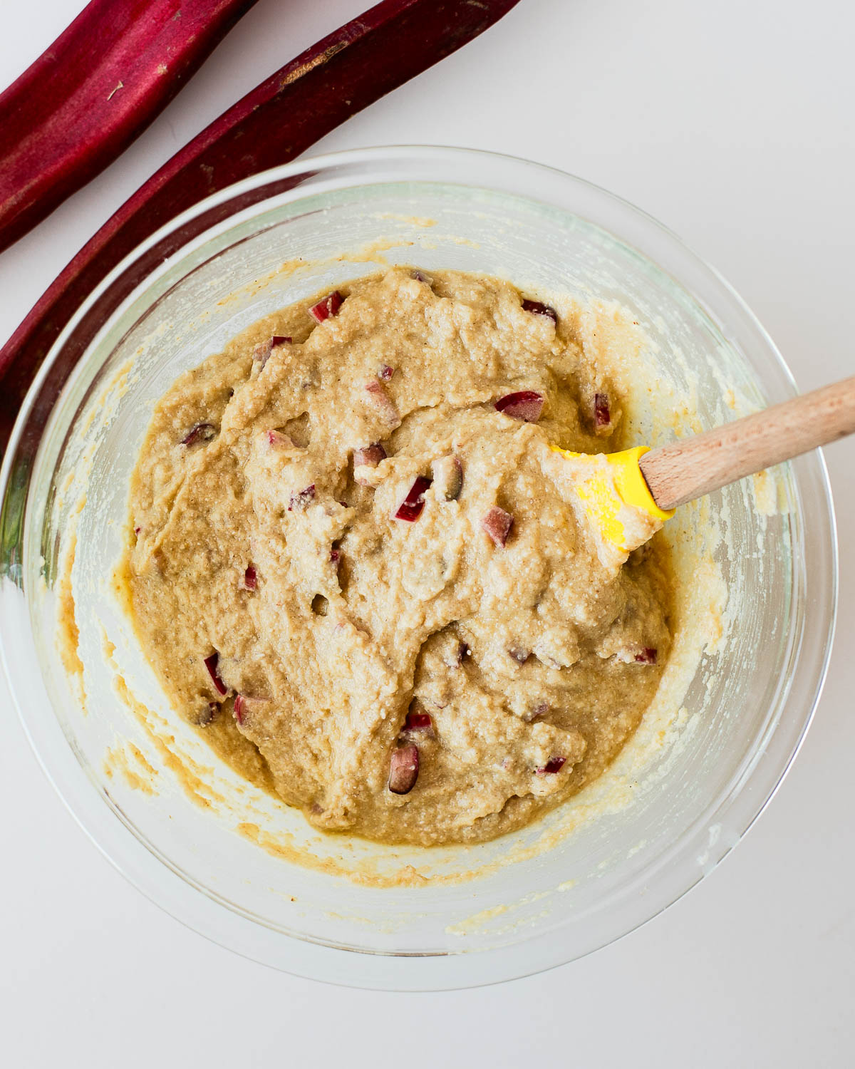 Mixing bowl of muffin batter with diced rhubarb being folded in with a spatula.