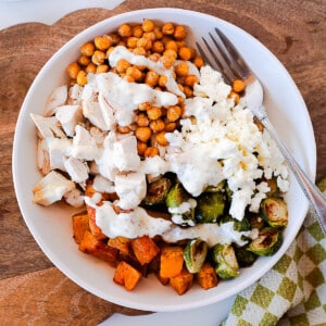 Close-up overhead view of a harvest chicken bowl with juicy rotisserie chicken, crispy chickpeas, roasted brussels sprouts, butternut squash, and feta cheese drizzled with creamy dressing.