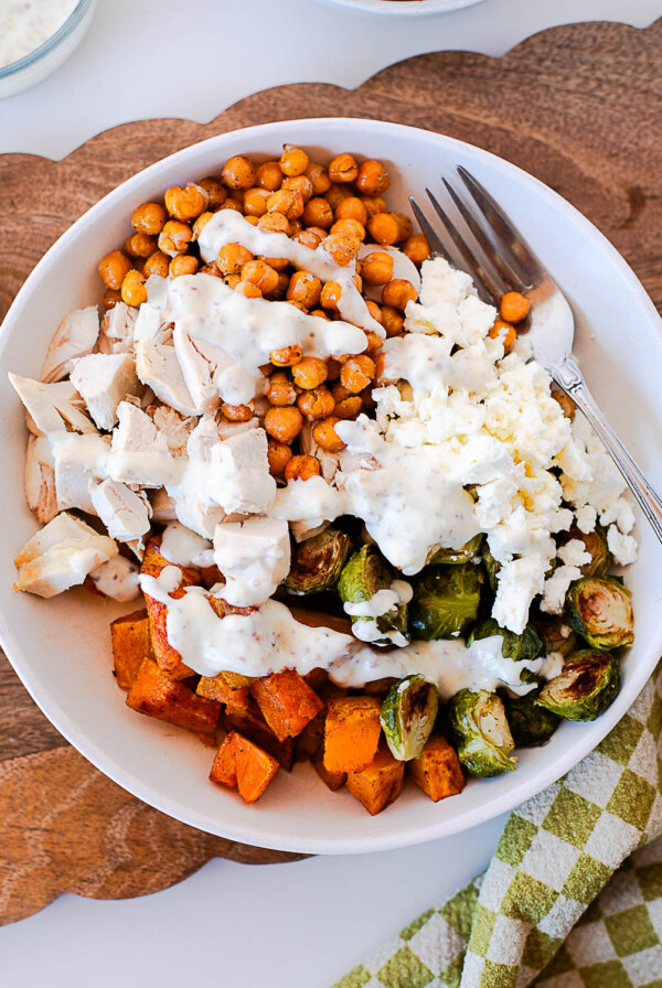 Close-up overhead view of a harvest chicken bowl with juicy rotisserie chicken, crispy chickpeas, roasted brussels sprouts, butternut squash, and feta cheese drizzled with creamy dressing.