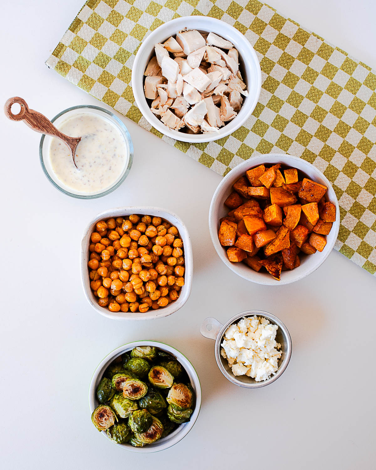 Flat lay of individual harvest bowl ingredients — bowls of roasted chickpeas, cubed chicken, feta cheese, roasted brussels sprouts, butternut squash, and a jar of creamy maple mustard dressing on a green checkered napkin.
