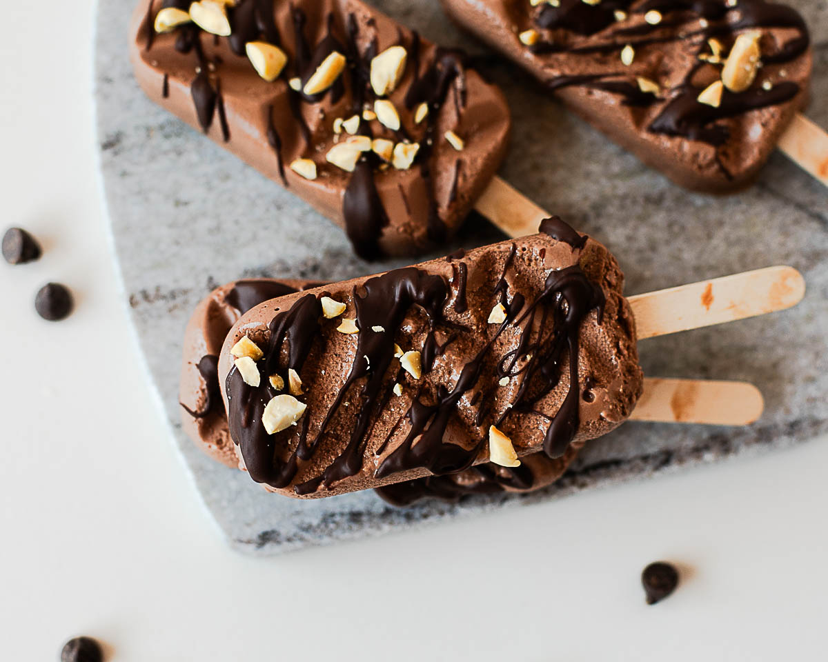Close-up of homemade chocolate protein popsicles finished with a dark chocolate drizzle and crunchy chopped peanuts for a healthy frozen dessert.