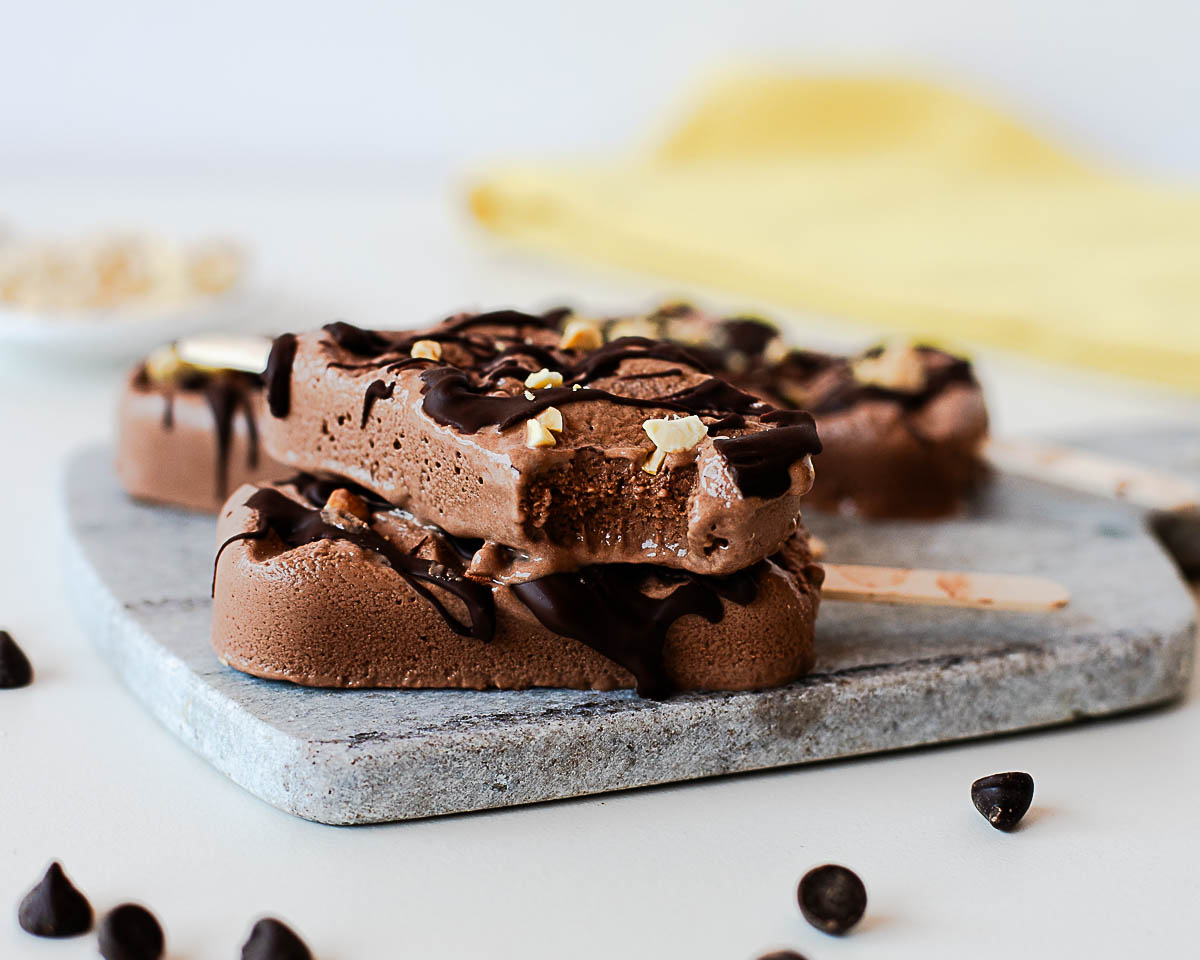 Chocolate protein popsicle with a bite taken out, showing the creamy fudgy center and topped with dark chocolate drizzle and chopped peanuts.