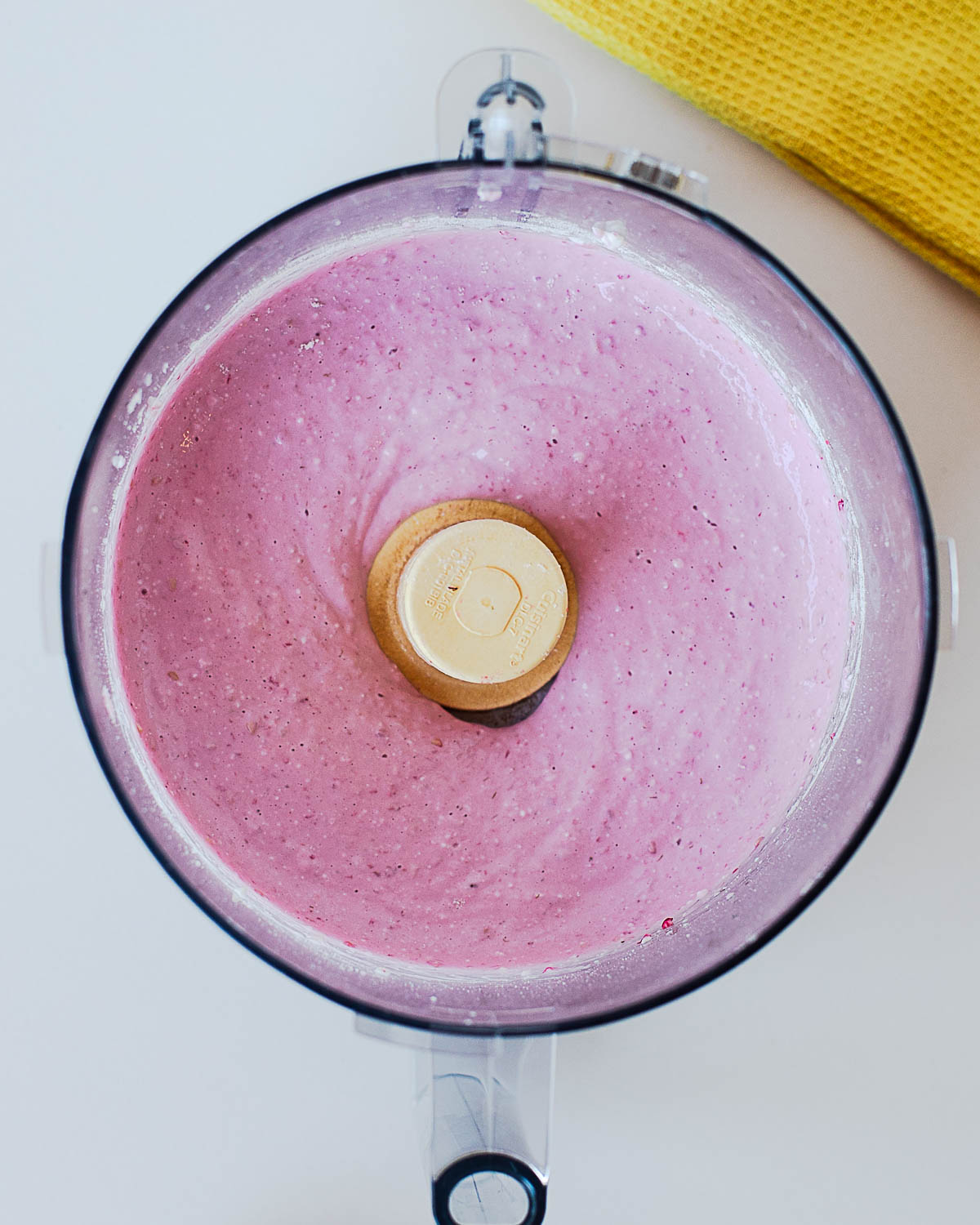 Top-down view of raspberry protein ice cream ingredients blended into a smooth, creamy pink mixture in a food processor before transferring to a Ninja Creami pint.
