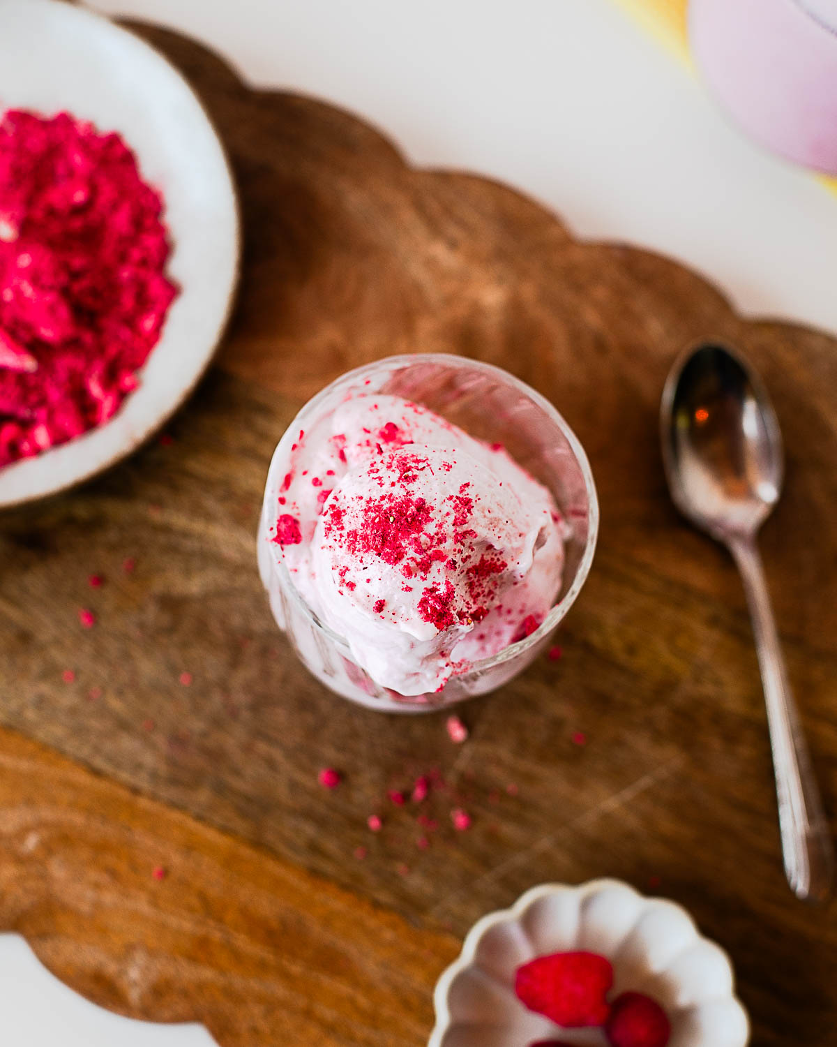 Overhead view of a scoop of raspberry protein ice cream in a glass dessert cup with freeze-dried raspberry topping on a wooden serving board.