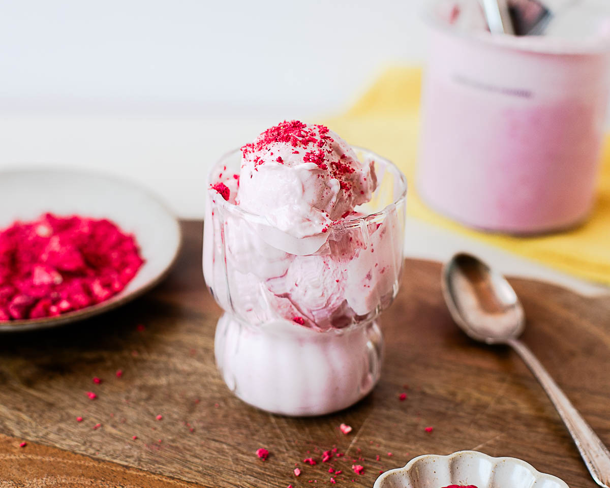 High-protein raspberry ice cream served in a glass dessert cup with freeze-dried raspberries, fresh berries, and a Ninja Creami pint in the background.